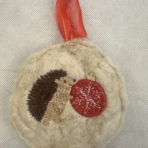 Cream wool ornament embroidered with a brown hedgehog holding a red Christmas bauble, finished with a red ribbon hanger.