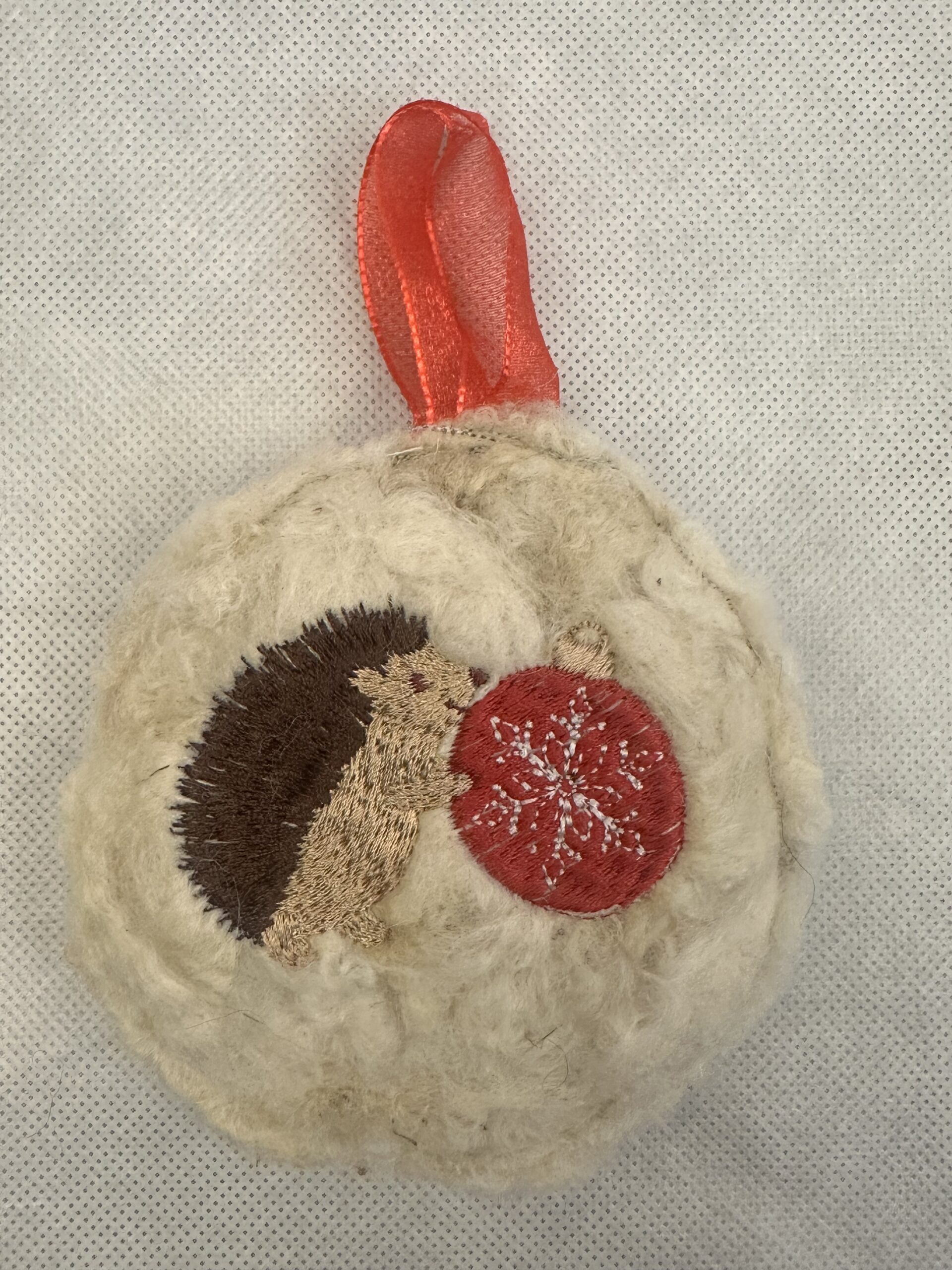 Cream wool ornament embroidered with a brown hedgehog holding a red Christmas bauble, finished with a red ribbon hanger.