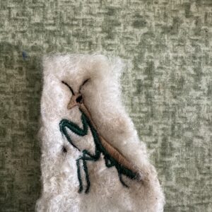 Small praying mantis with green legs, a brown body and dark antennae, embroidered on cream wool.