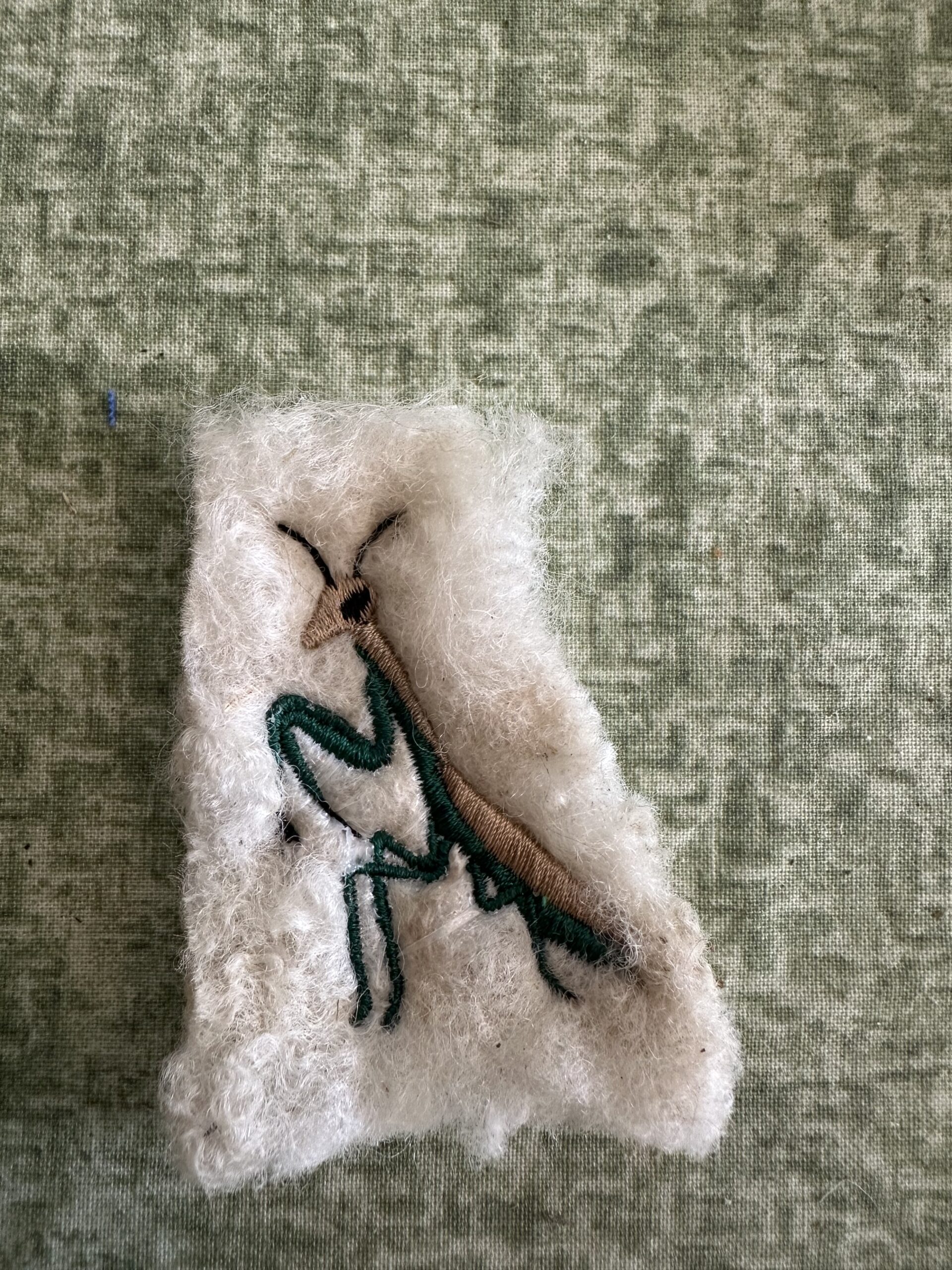 Small praying mantis with green legs, a brown body and dark antennae, embroidered on cream wool.