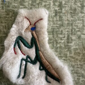 Large embroidered praying mantis with green legs, a brown body, blue head detail and reddish antennae on cream wool.