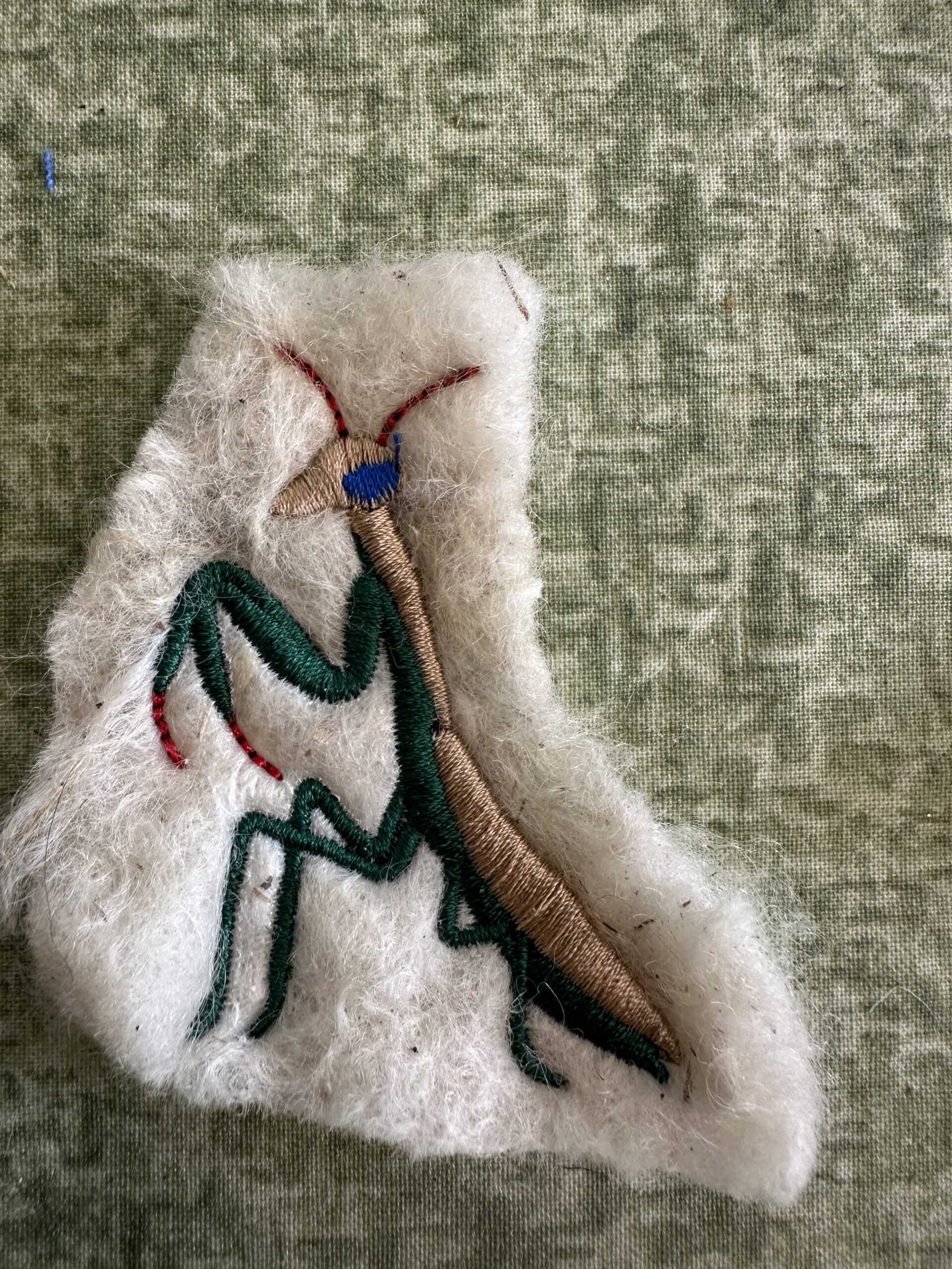 Large embroidered praying mantis with green legs, a brown body, blue head detail and reddish antennae on cream wool.